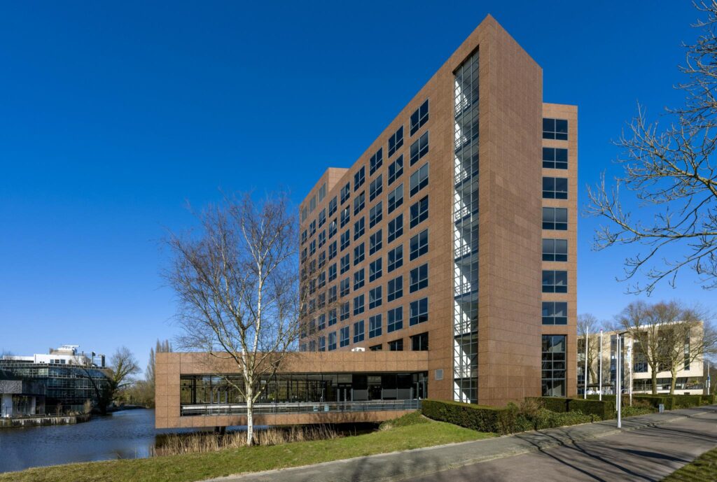 Modern kantoorgebouw aan de Prof. W.H. Keesomlaan met roodbruine gevel, grote ramen en een waterpartij op de voorgrond.
